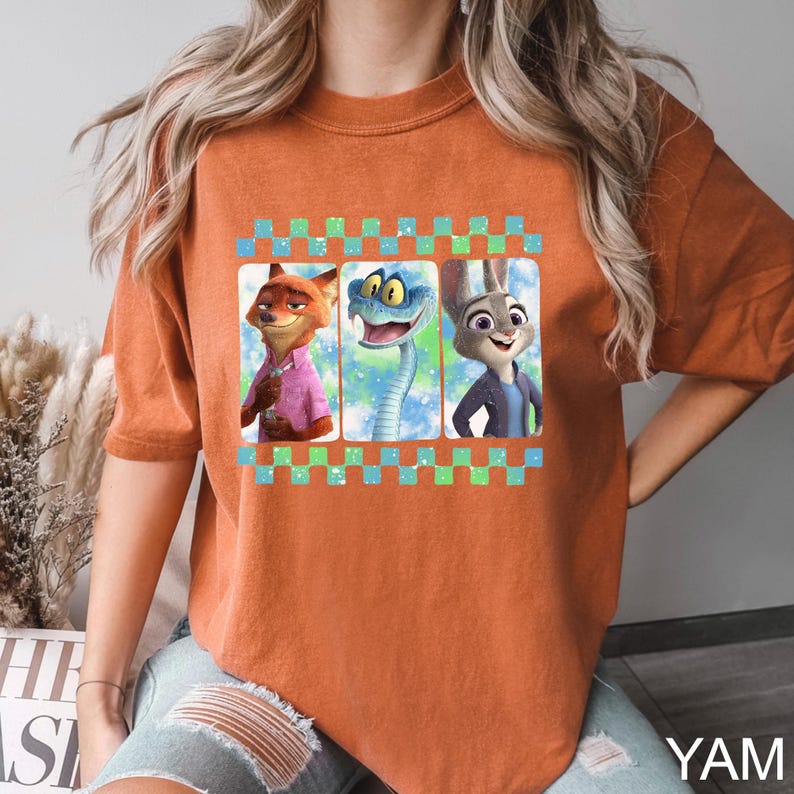 May include: Burnt orange t-shirt featuring a graphic of three cartoon characters: a fox, a snake, and a rabbit. The characters are set against a blue and green checkered background. The shirt has short sleeves and a relaxed fit. The text "YAM" is visible in the bottom right corner.
