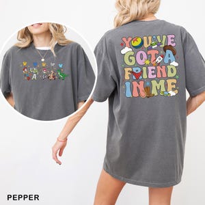 May include: Dark grey t-shirt featuring a colourful graphic on the back with the text "You've Got A Friend In Me." The front of the shirt has a smaller graphic of cartoon characters. The word "PEPPER" is printed at the bottom.