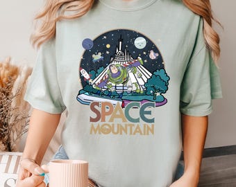 May include: A light blue t-shirt with a retro-style graphic of Buzz Lightyear riding Space Mountain at Disney World. The graphic features a colorful, stylized text that reads "Space Mountain".