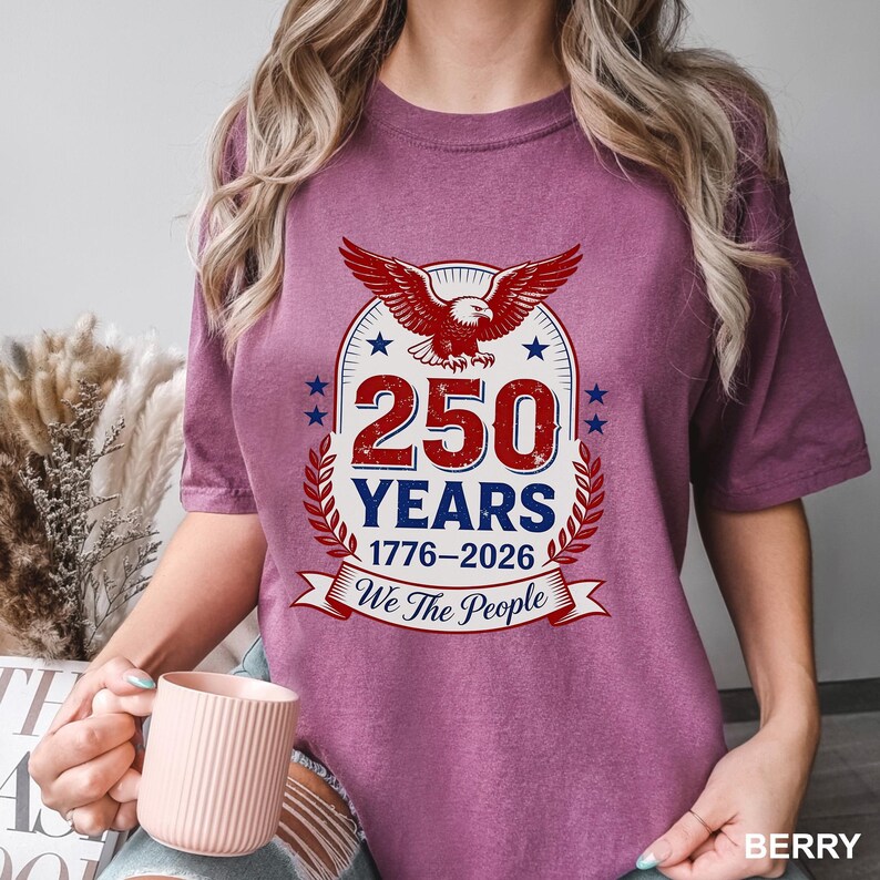 May include: Berry-colored t-shirt featuring a patriotic design. The graphic includes a red eagle, the text "250 YEARS 1776-2026", and the phrase "We The People". The design is framed by a laurel wreath and stars.