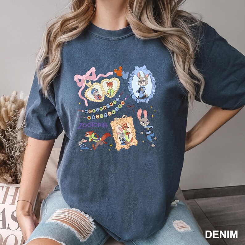 May include: Denim blue t-shirt featuring colorful illustrations of characters from the animated film "Zootopia." The design includes framed portraits, a pink bow, and the film's title. The shirt is made of a soft material.