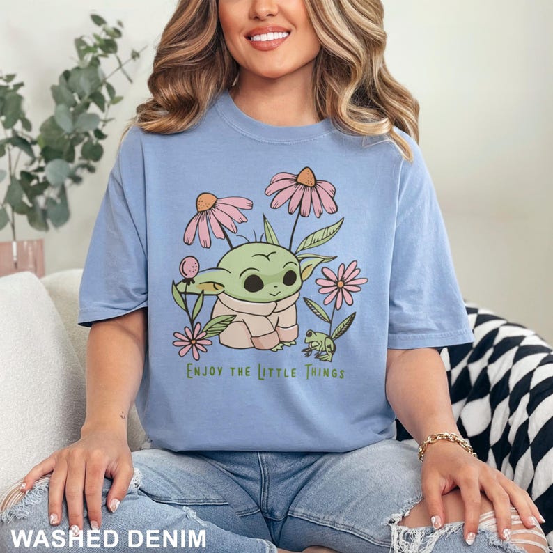 May include: A light blue t-shirt featuring a cartoon image of a green character with large ears and a frog. The shirt has the text "Enjoy the Little Things" printed on it.