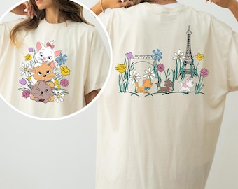 May include: Ivory-colored t-shirt featuring a colorful graphic of three cartoon cats surrounded by flowers on the front. The back of the shirt displays a scene with the Eiffel Tower and more cartoon cats. The word "IVORY" is printed at the bottom.