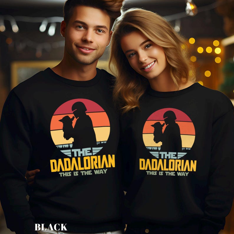 May include: A black crewneck sweatshirt featuring a graphic of a silhouette of a helmeted figure holding a small creature against a sunset background. The text reads THE DADALORIAN THIS IS THE WAY in yellow and white lettering on the front.