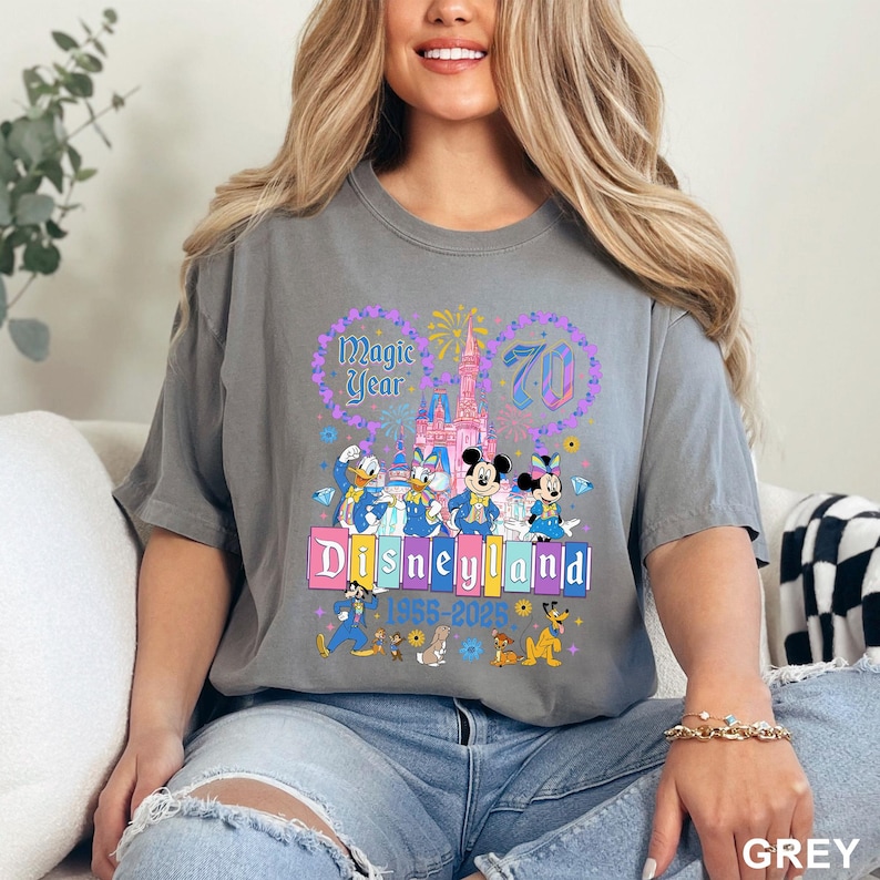 May include: A grey t-shirt featuring a colorful Disneyland graphic. The design includes the text "Magic Year 70" and "Disneyland 1955-2025" with images of Disney characters like Mickey and Minnie Mouse, Donald Duck, and Goofy.