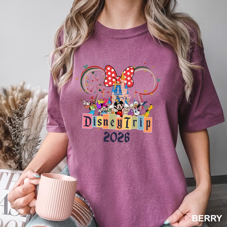 May include: Berry-colored t-shirt featuring a colorful Disney Trip 2026 graphic. The design includes Mickey Mouse, Minnie Mouse, and other Disney characters, with a castle and a polka-dot bow. The shirt is a short-sleeved crew neck.