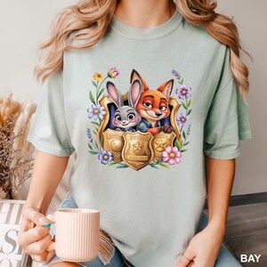 May include: A pale green t-shirt with a vibrant image of Judy Hopps and Nick Wilde from Zootopia, surrounded by flowers and police badges. The tee has a round neck and short sleeves. The word "BAY" is printed on the bottom right.