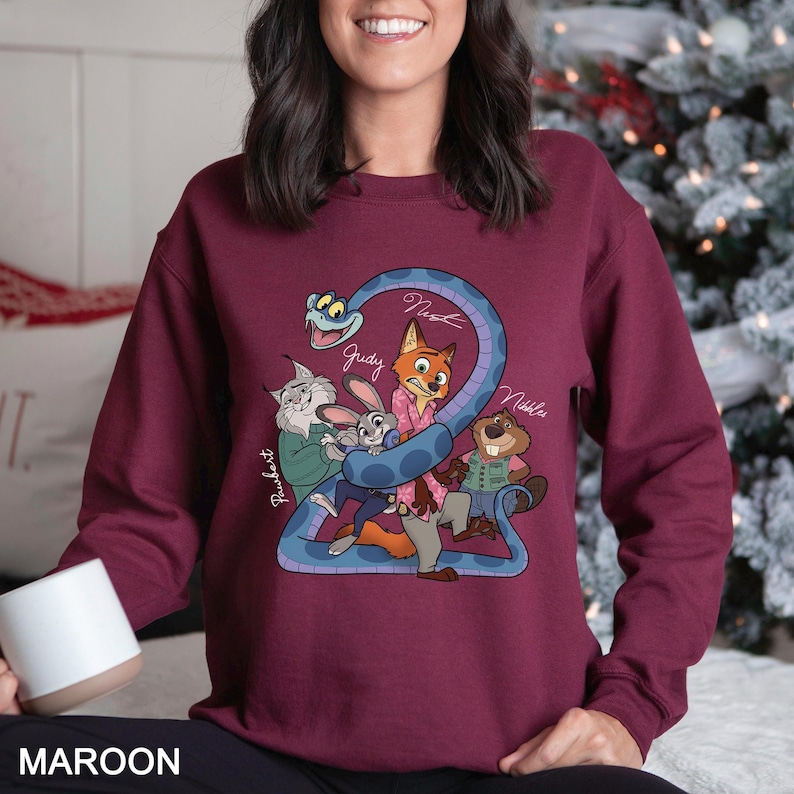 Puede incluir: Sudadera granate con un gr&aacute;fico de dibujos animados de personajes de pel&iacute;culas. El gr&aacute;fico incluye una serpiente azul con el n&uacute;mero 2 y los nombres Judy, Nick y Nibbles. La palabra "MAROON" est&aacute; impresa en la parte inferior.