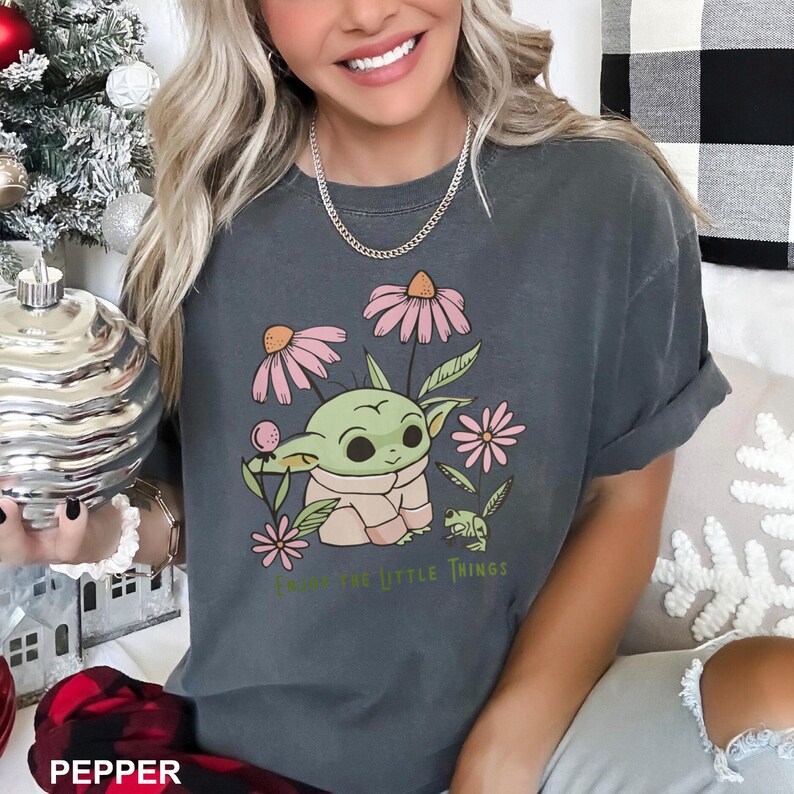 May include: A dark grey t-shirt featuring a cartoon image of a green alien character surrounded by pink flowers and a green frog. The text "Enjoy the little things" is printed below the image.