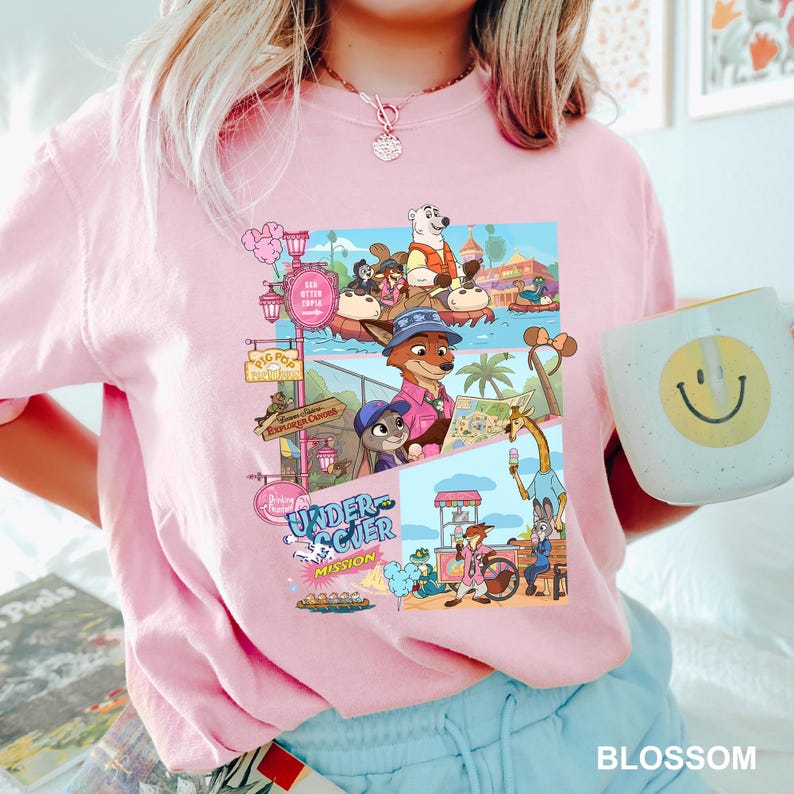 May include: Pale pink t-shirt with a vibrant graphic depicting cartoon characters in a theme park. The design includes the text "Under Scier Mission" and other signs. A white mug with a yellow smiley face is also visible.