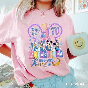May include: Pink t-shirt featuring a Disneyland 70th anniversary design. The graphic includes Mickey and Minnie Mouse, Donald and Daisy Duck, and the Disneyland castle. Text reads "Magic Year" and "Disneyland 1955-2025".
