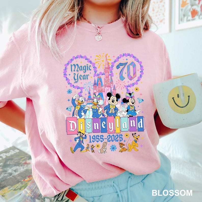 May include: Pink t-shirt featuring a Disneyland 70th anniversary design. The graphic includes Mickey and Minnie Mouse, Donald and Daisy Duck, and the Disneyland castle. Text reads "Magic Year" and "Disneyland 1955-2025".