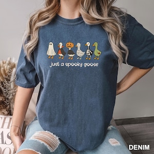 May include: A denim blue t-shirt featuring a line of cartoon geese dressed in Halloween costumes. The text "just a spooky goose" is printed below the geese. The shirt is a comfortable, casual style.