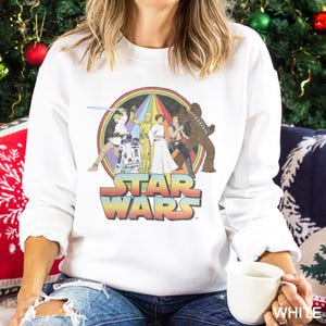K&ouml;nnte beinhalten: Wei&szlig;er Pullover mit einem Regenbogen-Motiv von Star Wars-Charakteren, darunter Luke Skywalker, Prinzessin Leia, Chewbacca, R2-D2 und Darth Vader. Der Text "Star Wars" ist unter dem Motiv gedruckt.