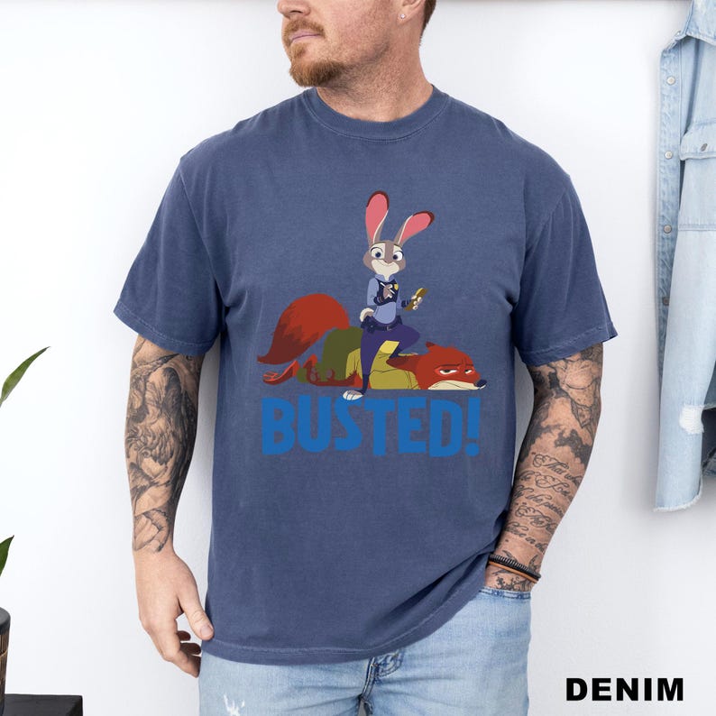 May include: A denim blue t-shirt featuring a cartoon illustration of a rabbit police officer standing on a fox, with the word "BUSTED!" in blue block letters. The shirt has short sleeves and a crew neck.