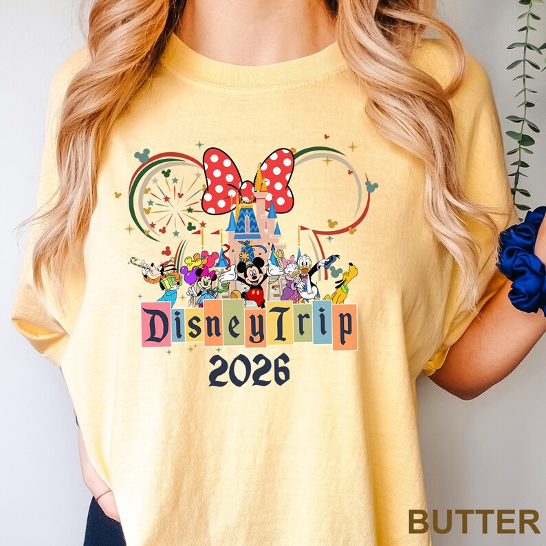 May include: Butter yellow t-shirt featuring a Disney Trip design. The design includes a castle, Minnie Mouse bow, and various Disney characters. The text "DisneyTrip 2026" is prominently displayed.