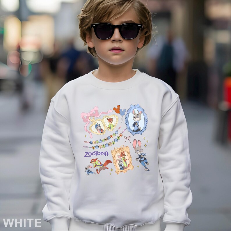May include: A white sweatshirt featuring colorful cartoon illustrations from the movie Zootopia. The design includes framed character portraits of Judy Hopps, Nick Wilde, and other characters. The word "Zootopia" is printed in purple.