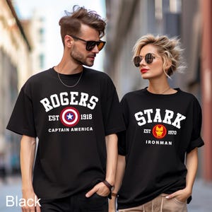 May include: Two black t-shirts with a vintage college style design. The first shirt says "Rogers Est 1918 Captain America" with a red, white, and blue Captain America shield. The second shirt says "Stark Est 1970 Ironman" with a red and gold Ironman mask.
