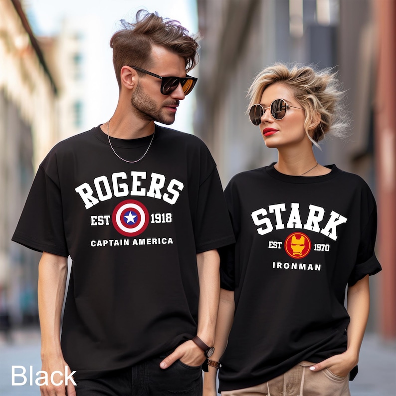 May include: Two black t-shirts with a vintage college style design. The first shirt says "Rogers Est 1918 Captain America" with a red, white, and blue Captain America shield. The second shirt says "Stark Est 1970 Ironman" with a red and gold Ironman mask.