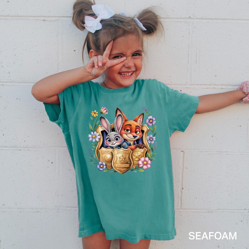 May include: A teal t-shirt with a graphic of Judy Hopps and Nick Wilde from Zootropolis, framed by flowers and a gold badge. The shirt has short sleeves and the word "SEAFOAM" printed at the bottom.