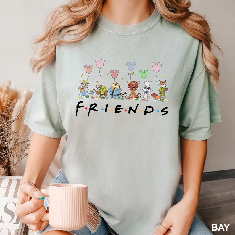 May include: A light green t-shirt featuring the word "FRIENDS" in a colorful font. Above the word are cartoon characters holding Mickey Mouse-shaped balloons. The shirt is a soft, comfortable material.