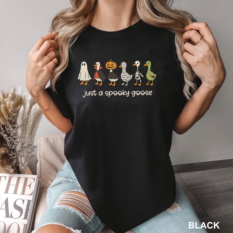 May include: Black t-shirt with a Halloween-themed design. It features a row of cartoon geese in costumes, including a ghost, vampire, pumpkin, mummy, skeleton, and a green goose. The text "just a spooky goose" is printed below.