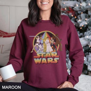 K&ouml;nnte beinhalten: Ein maroonfarbener Pullover mit einem Vintage-Star-Wars-Motiv, das Figuren wie Luke Skywalker, Prinzessin Leia, Han Solo, Chewbacca und R2-D2 zeigt. Das Motiv ist von einem regenbogenfarbenen Kreis mit dem Text "Star Wars" in gelber und orangefarbener Schrift umgeben.
