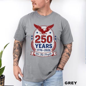 May include: Grey t-shirt featuring a patriotic design. The design includes a red eagle, the text "250 YEARS 1776-2026", and the phrase "We the People". The shirt is a short-sleeved crew neck.