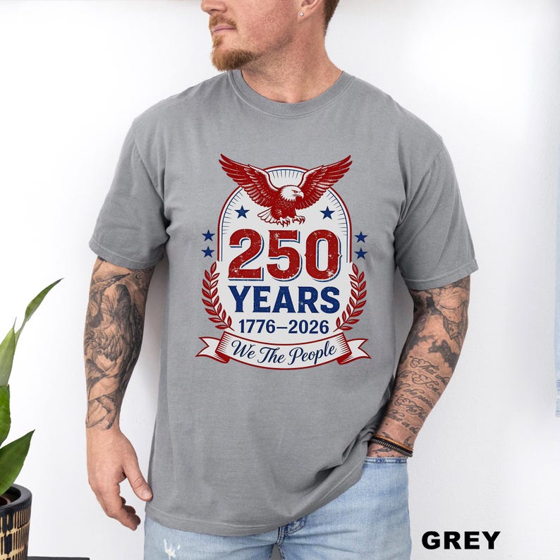 May include: Grey t-shirt featuring a patriotic design. The design includes a red eagle, the text "250 YEARS 1776-2026", and the phrase "We the People". The shirt is a short-sleeved crew neck.