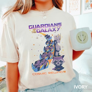 May include: Ivory-colored t-shirt featuring a graphic of Guardians of the Galaxy characters. The design includes the text "GUARDIANS OF THE GALAXY" and "COSMIC REWIND" in purple and orange. A white mug with a smiley face is held in the image.