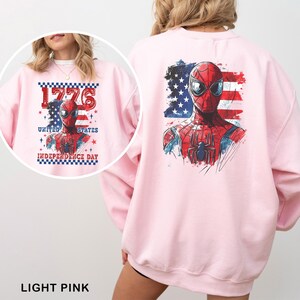 May include: A light pink sweatshirt featuring a graphic of Spider-Man against a distressed American flag background. The design includes the text 1776 UNITED STATES INDEPENDENCE DAY with a checkered border and small star accents in red and blue.