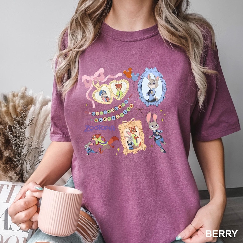 May include: Berry-colored t-shirt featuring colorful illustrations of characters from the animated movie "Zootopia." The design includes framed portraits of the characters, a string of lights, and the movie's logo. The shirt is a comfortable, casual style.