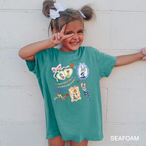 May include: A teal-colored t-shirt featuring framed cartoon characters from Zootopia. The shirt has a pink bow and the word "Zootopia" in a script font. The bottom right corner of the shirt has the word "SEAFOAM".