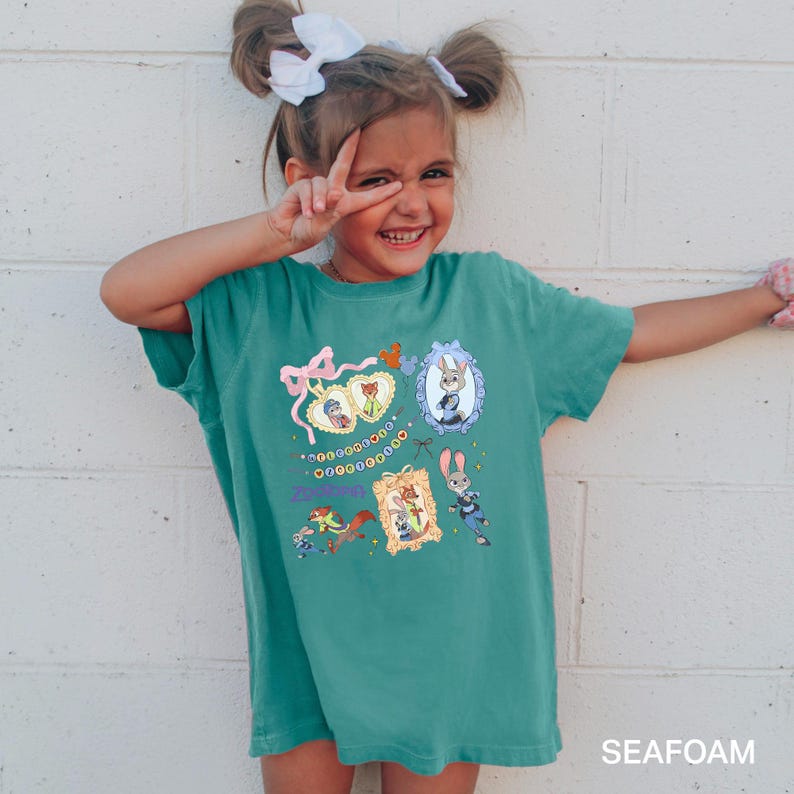 May include: A teal-colored t-shirt featuring framed cartoon characters from Zootopia. The shirt has a pink bow and the word "Zootopia" in a script font. The bottom right corner of the shirt has the word "SEAFOAM".