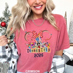 May include: Crimson-colored t-shirt featuring a Disney Trip design. The design includes Mickey Mouse, Minnie Mouse, and other Disney characters, a castle, and the text "Disney Trip 2026". The shirt has short sleeves.