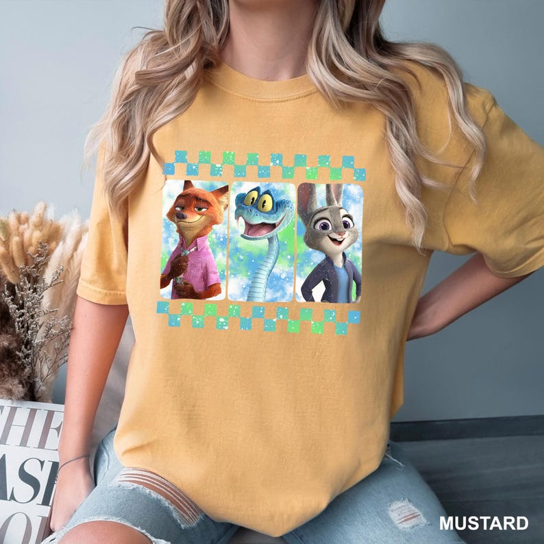 May include: Mustard-colored t-shirt featuring a graphic of three characters from the animated film Zootopia: Nick Wilde the fox, a snake, and Judy Hopps the rabbit. The characters are in rectangular frames with a blue and green checkered border.
