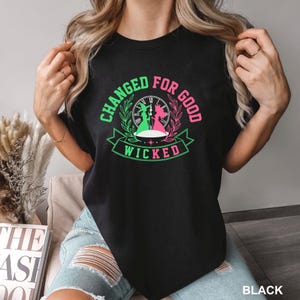 May include: A black t-shirt featuring a graphic print with the text "CHANGED FOR GOOD" in green and pink, and "WICKED" in green. The design includes a clock face, silhouettes of two figures, and decorative leaf motifs in green and pink.