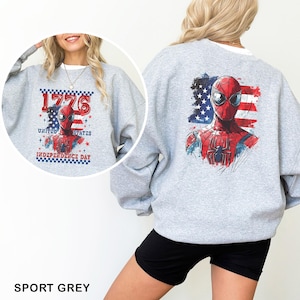 May include: A light grey crewneck sweatshirt featuring a graphic of the superhero Spider-Man centred on the back, layered over a distressed American flag design. The front includes the same graphic with the text 1776 UNITED STATES INDEPENDENCE DAY.