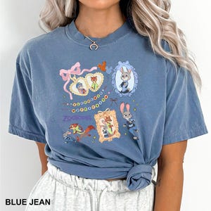 May include: Blue jean-colored t-shirt featuring a colorful graphic of characters and imagery from the animated film "Zootopia." The design includes framed portraits, bows, and the film's title. The shirt is a casual, short-sleeved style.