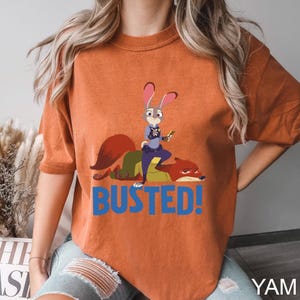 May include: Orange t-shirt featuring a cartoon illustration of a rabbit police officer standing over a fox, with the word "BUSTED!" in blue. The rabbit is holding a carrot. The shirt is a solid orange color.