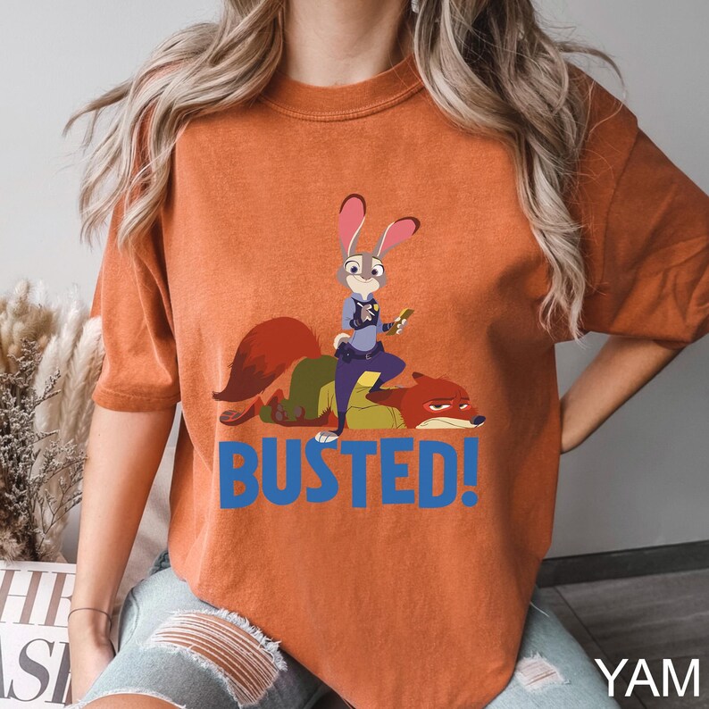May include: Orange t-shirt featuring a cartoon illustration of a rabbit police officer standing over a fox, with the word "BUSTED!" in blue. The rabbit is holding a carrot. The shirt is a solid orange color.