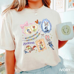 May include: Ivory-colored t-shirt featuring colorful Zootopia character illustrations. The design includes framed portraits of Judy Hopps and Nick Wilde, along with the word "Zootopia" and the phrase "Welcome to Zootopia". A white mug with a smiley face is held in the image.