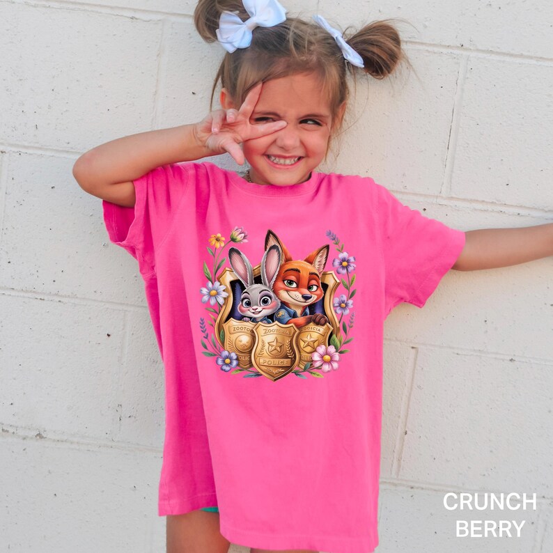 May include: A vibrant pink t-shirt with a graphic of Judy Hopps and Nick Wilde from Zootopia within a police badge, framed by flowers. The shirt is labelled "Crunch Berry" at the bottom right.