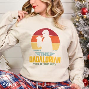 May include: A sand-coloured crewneck sweatshirt featuring a retro-style graphic of a helmeted figure holding a small creature inside a sunset circle. The text reads THE DADALORIAN THIS IS THE WAY in yellow and teal lettering with a wing-like design.