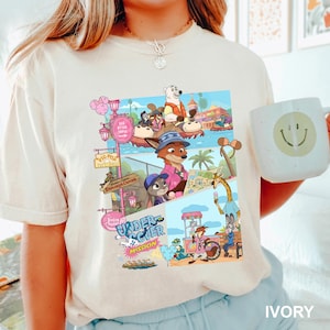 May include: Cream-coloured t-shirt with a vibrant graphic of cartoon characters in a theme park. The graphic includes the text "Undercover Mission" and "Explorer Candies". A white mug with a smiley face is held in the image.