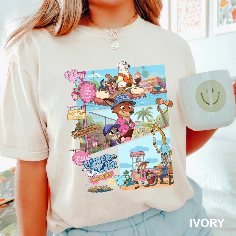 May include: Cream-coloured t-shirt with a vibrant graphic of cartoon characters in a theme park. The graphic includes the text "Undercover Mission" and "Explorer Candies". A white mug with a smiley face is held in the image.
