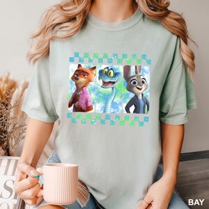 May include: A light green t-shirt featuring a graphic of three cartoon characters: a fox, a snake, and a rabbit. The characters are set against a colorful background within a checkered border. The shirt is a light green color.