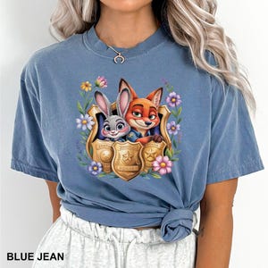 May include: A blue jean coloured t-shirt with a colourful graphic of Judy Hopps and Nick Wilde from Zootopia, framed by flowers and police badges. The text "Zootopia Police" is visible on the badges. The shirt is a comfortable, casual style.