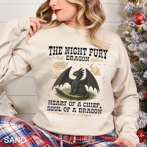 Puede incluir: Una sudadera color arena con un gr&aacute;fico de drag&oacute;n negro y el texto "THE NIGHT FURY DRAGON". El dise&ntilde;o incluye las frases "Warning Never Engage This Dragon" y "HEART OF A CHIEF, SOUL OF A DRAGON."