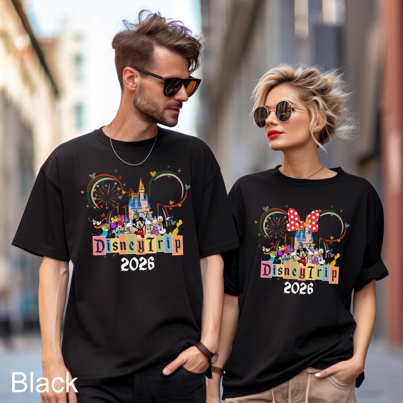 May include: Black t-shirts with a colorful Disney Trip 2026 design. The design includes a castle, characters, and a Ferris wheel. The shirts are worn by two people, one with sunglasses.
